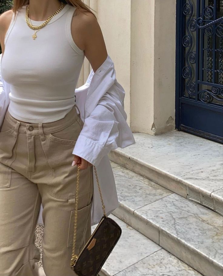 A young lady model styling a white button down shirt with tank top and beige color cargo pants look so pretty to hold a clutch in outdoors.