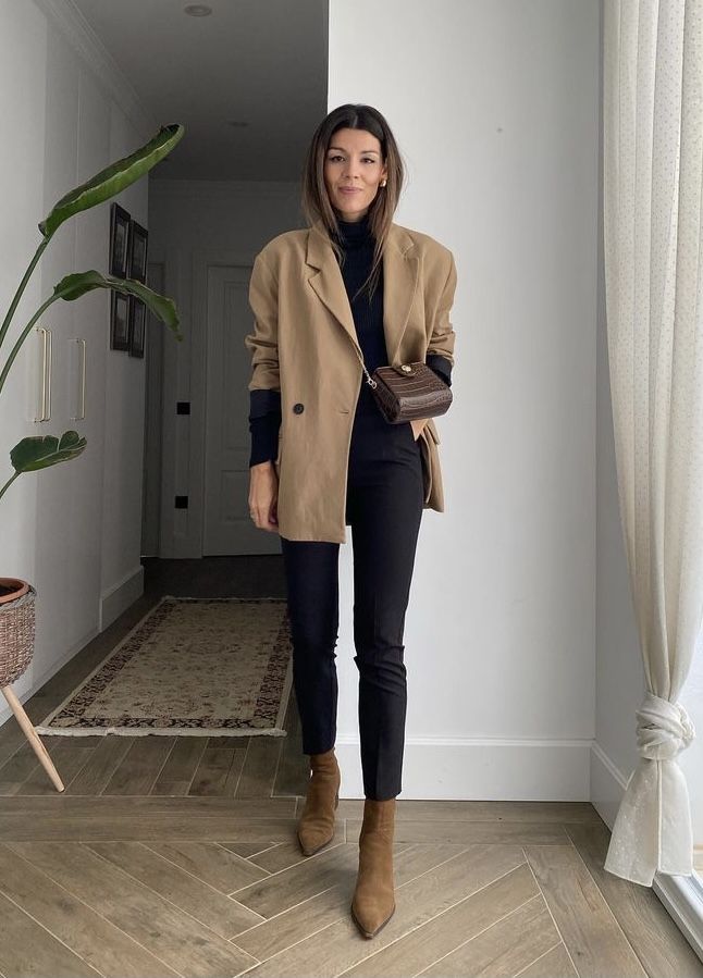 A young woman posing in the house to style a brown coat with black jeans and brown ankle boots. 