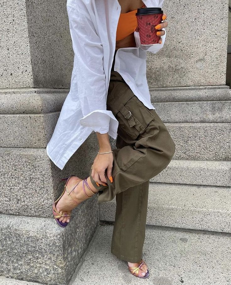 Styling white oversized shirt with vest or cargo pants and high heels a young girl holding coffee mug in her hand in outdoors. 